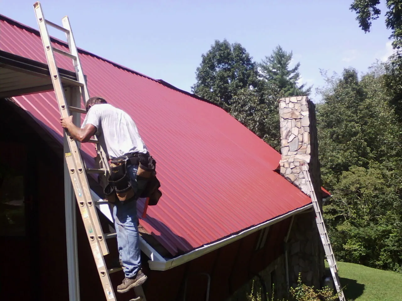 Roofer on a ladder beside a red metal roof and stone chimney — Soffit & Fascia Repair by Roofing Brothers in New Tripoli